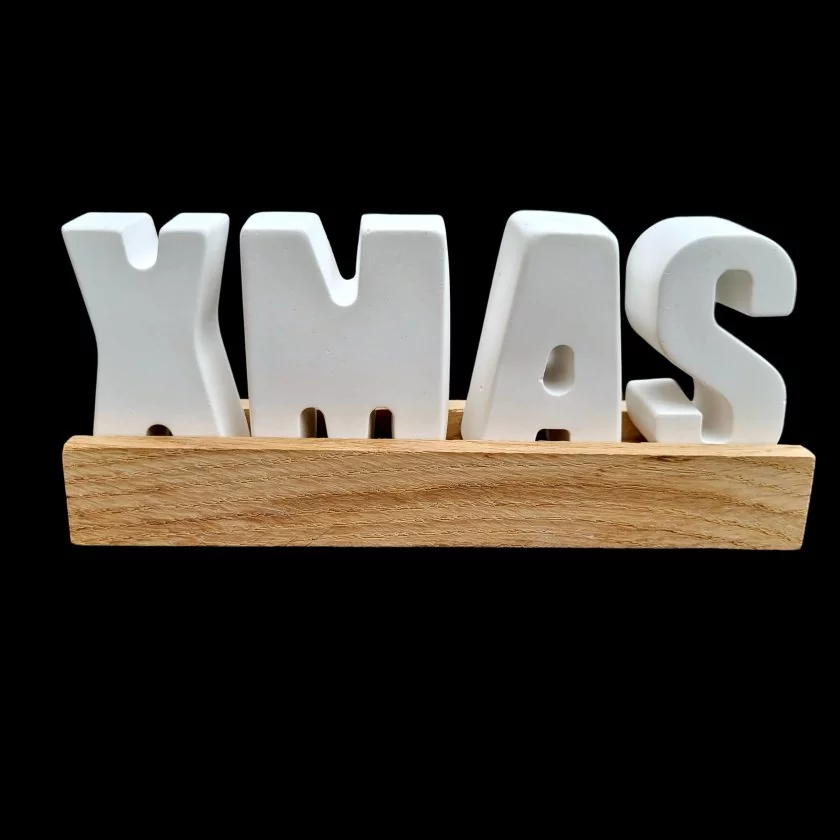Weißer Buchstaben-Schriftzug "XMAS" aus Raysin auf Holzbox vor schwarzem Hintergrund